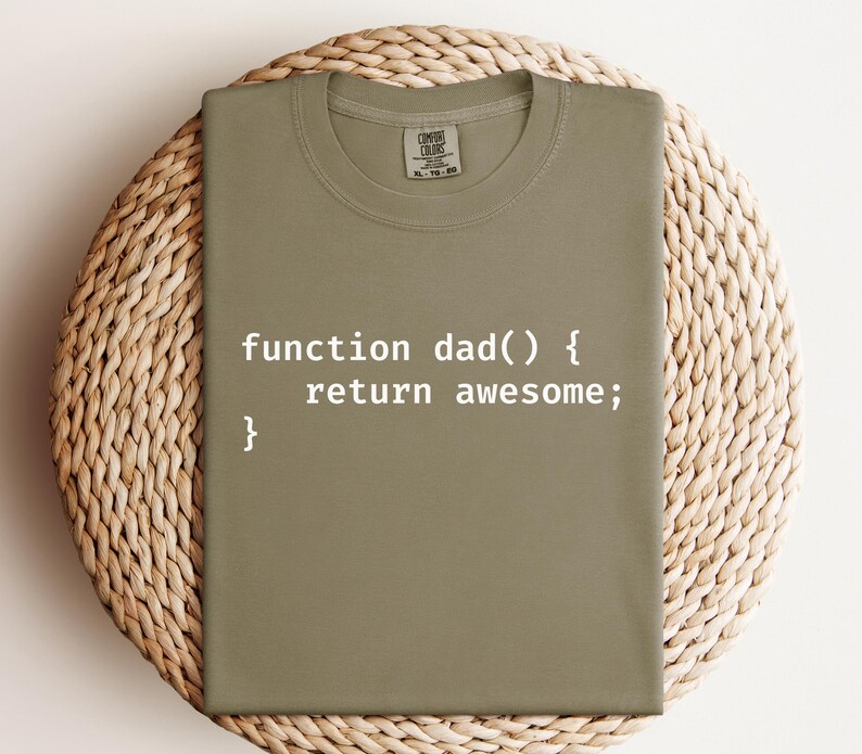K&ouml;nnte beinhalten: Olivgr&uuml;nes T-Shirt mit dem wei&szlig;en Text "function dad() { return awesome; }". Das T-Shirt wird auf einer gewebten, kreisf&ouml;rmigen Oberfl&auml;che pr&auml;sentiert.