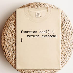 K&ouml;nnte beinhalten: Beiges T-Shirt mit dem schwarzen Text "function dad() { return awesome; }". Das Shirt liegt auf einer gewebten, kreisf&ouml;rmigen Oberfl&auml;che. Das Shirt hat einen Rundhalsausschnitt und ein Etikett am Ausschnitt.