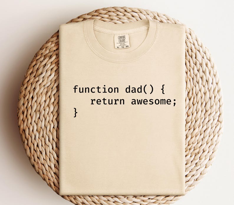 K&ouml;nnte beinhalten: Beiges T-Shirt mit dem schwarzen Text "function dad() { return awesome; }". Das Shirt liegt auf einer gewebten, kreisf&ouml;rmigen Oberfl&auml;che. Das Shirt hat einen Rundhalsausschnitt und ein Etikett am Ausschnitt.