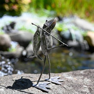 May include: A metal frog sculpture holding a fish in its hand. The frog is standing on a rock with a stream in the background.