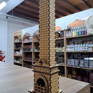 Op de afbeelding: Een houten model van een open haard met een hoge schoorsteen. De open haard is gemaakt van lichtbruin hout en heeft een zwart metalen rooster. De schoorsteen is gemaakt van gestapelde bakstenen en heeft een klein decoratief detail bovenaan.