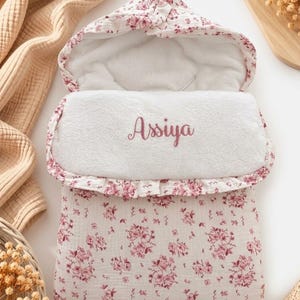 Peut inclure: Une gigoteuse pour bébé avec capuche, présentant une base en tissu blanc avec un motif floral rose. L'intérieur est doublé de tissu blanc et le prénom Assiya est brodé en fil rose sur le rabat avant, qui possède un bord à volants.
