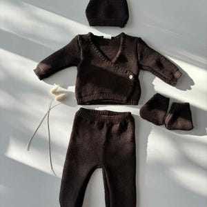 K&ouml;nnte beinhalten: Ein dunkelbraunes Baby-Kleidungsset, bestehend aus einem Wickel-Cardigan, einer Hose, einer M&uuml;tze und Babyschuhen. Das Strickset ist aus weichem Material gefertigt und wird auf einer wei&szlig;en Oberfl&auml;che pr&auml;sentiert. Der Cardigan hat einen Knopfverschluss.