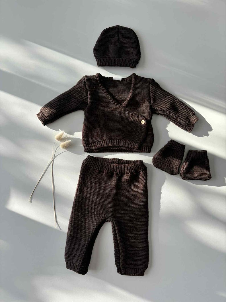 K&ouml;nnte beinhalten: Ein dunkelbraunes Baby-Kleidungsset, bestehend aus einem Wickel-Cardigan, einer Hose, einer M&uuml;tze und Babyschuhen. Das Strickset ist aus weichem Material gefertigt und wird auf einer wei&szlig;en Oberfl&auml;che pr&auml;sentiert. Der Cardigan hat einen Knopfverschluss.