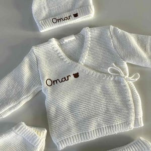 K&ouml;nnte beinhalten: Ein wei&szlig;es Strick-Baby-Outfit-Set, bestehend aus einer M&uuml;tze, einem Wickel-Cardigan und Shorts. Der Name "Omar" ist in Braun auf die M&uuml;tze und den Cardigan gestickt. Das Set ist aus einem strukturierten Strickstoff gefertigt.