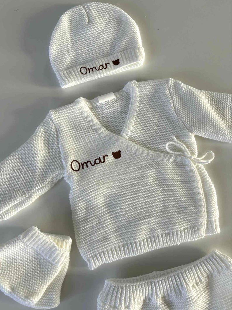 K&ouml;nnte beinhalten: Ein wei&szlig;es Strick-Baby-Outfit-Set, bestehend aus einer M&uuml;tze, einem Wickel-Cardigan und Shorts. Der Name "Omar" ist in Braun auf die M&uuml;tze und den Cardigan gestickt. Das Set ist aus einem strukturierten Strickstoff gefertigt.