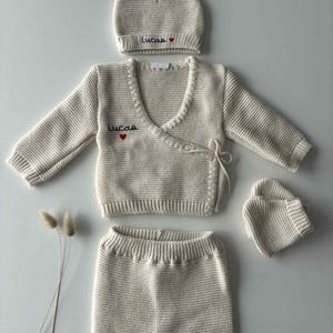 K&ouml;nnte beinhalten: Cremefarbenes Baby-Kleidungsset, bestehend aus M&uuml;tze, Wickelpullover, Hose und F&auml;ustlingen. M&uuml;tze und Pullover sind mit dem Namen "Lucas" und einem roten Herz bestickt. Das Set ist aus Strickmaterial gefertigt.