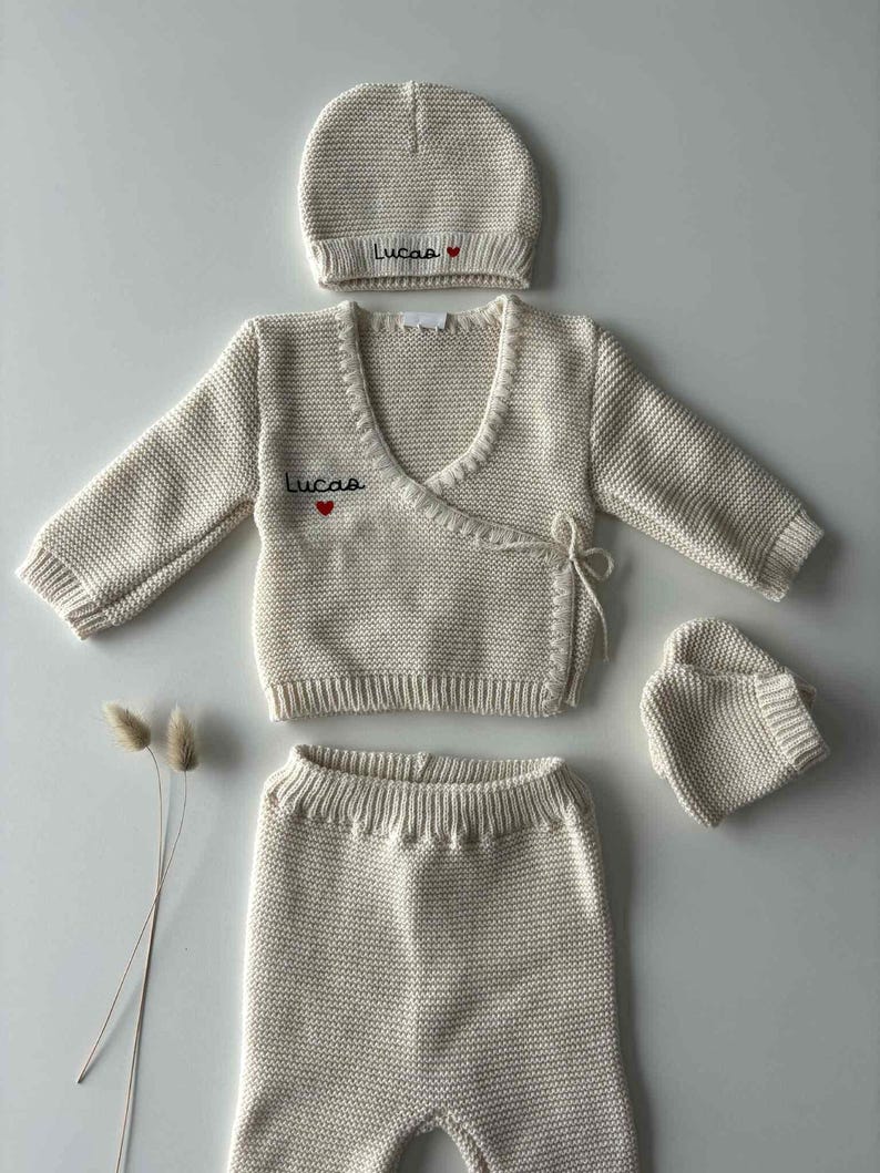 K&ouml;nnte beinhalten: Cremefarbenes Baby-Kleidungsset, bestehend aus M&uuml;tze, Wickelpullover, Hose und F&auml;ustlingen. M&uuml;tze und Pullover sind mit dem Namen "Lucas" und einem roten Herz bestickt. Das Set ist aus Strickmaterial gefertigt.