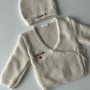 K&ouml;nnte beinhalten: Cremefarbenes Strick-Babyset, bestehend aus einem Wickel-Cardigan und einer M&uuml;tze. Der Name "Lucas" ist in Schwarz mit einem roten Herz gestickt. Der Cardigan hat lange &Auml;rmel und Bindeb&auml;nder.