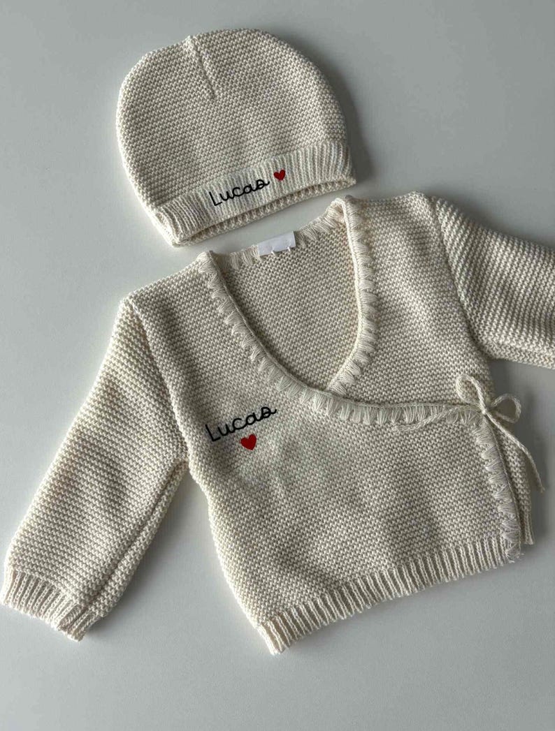 K&ouml;nnte beinhalten: Cremefarbenes Strick-Babyset, bestehend aus einem Wickel-Cardigan und einer M&uuml;tze. Der Name "Lucas" ist in Schwarz mit einem roten Herz gestickt. Der Cardigan hat lange &Auml;rmel und Bindeb&auml;nder.