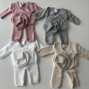 K&ouml;nnte beinhalten: Vier Baby-Outfits in Rosa, Grau, Wei&szlig; und Beige. Jedes Set enth&auml;lt einen Wickelpullover, eine Hose und eine passende M&uuml;tze. Das Strickmaterial ist weich und wirkt bequem f&uuml;r Neugeborene.