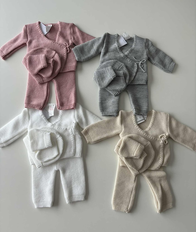 K&ouml;nnte beinhalten: Vier Baby-Outfits in Rosa, Grau, Wei&szlig; und Beige. Jedes Set enth&auml;lt einen Wickelpullover, eine Hose und eine passende M&uuml;tze. Das Strickmaterial ist weich und wirkt bequem f&uuml;r Neugeborene.