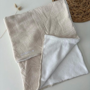 Peut inclure: Une couverture pour bébé en mousseline beige avec un intérieur en peluche blanche. La couverture est pliée, révélant la doublure blanche douce. Le nom "Liam" est brodé en écriture blanche avec un petit cœur.