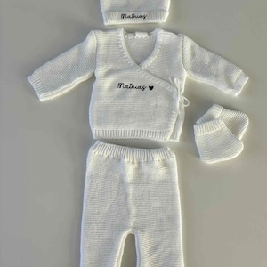 K&ouml;nnte beinhalten: Ein wei&szlig;es Strick-Baby-Outfit-Set, bestehend aus M&uuml;tze, Wickeloberteil, Hose und Babyschuhen. Der Name "Mathias" ist auf M&uuml;tze und Oberteil gestickt. Das Oberteil hat auch ein kleines Herzsymbol. Das Set ist aus weichem, strukturiertem Strick gefertigt.