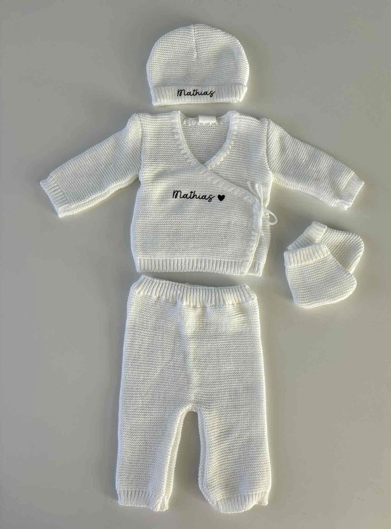 K&ouml;nnte beinhalten: Ein wei&szlig;es Strick-Baby-Outfit-Set, bestehend aus M&uuml;tze, Wickeloberteil, Hose und Babyschuhen. Der Name "Mathias" ist auf M&uuml;tze und Oberteil gestickt. Das Oberteil hat auch ein kleines Herzsymbol. Das Set ist aus weichem, strukturiertem Strick gefertigt.