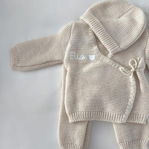 K&ouml;nnte beinhalten: Beige gestricktes Baby-Outfit-Set, bestehend aus einem Wickel-Cardigan, einer Hose und einer M&uuml;tze. Der Cardigan ist mit dem Namen "Elio" in Wei&szlig; bestickt. Das Set ist aus einem weichen, strukturierten Strickstoff gefertigt.