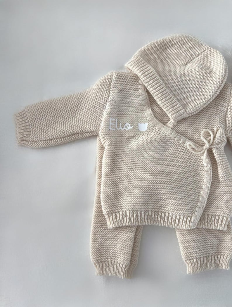 K&ouml;nnte beinhalten: Beige gestricktes Baby-Outfit-Set, bestehend aus einem Wickel-Cardigan, einer Hose und einer M&uuml;tze. Der Cardigan ist mit dem Namen "Elio" in Wei&szlig; bestickt. Das Set ist aus einem weichen, strukturierten Strickstoff gefertigt.