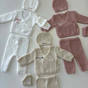 K&ouml;nnte beinhalten: Mehrere Baby-Kleidungssets in Creme, Rosa und Wei&szlig;. Jedes Set enth&auml;lt einen Wickelpullover, eine Hose und eine M&uuml;tze. Einige Sets haben Namen wie "Alma", "Roxane" und "Stan" aufgestickt.