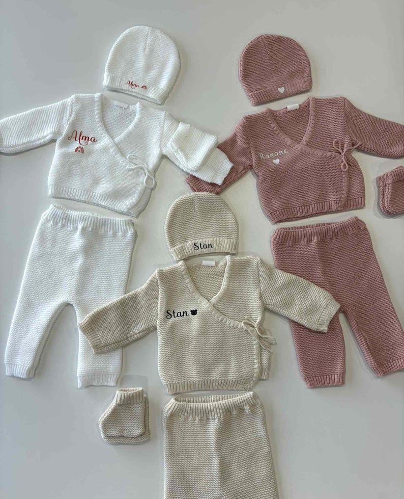 K&ouml;nnte beinhalten: Mehrere Baby-Kleidungssets in Creme, Rosa und Wei&szlig;. Jedes Set enth&auml;lt einen Wickelpullover, eine Hose und eine M&uuml;tze. Einige Sets haben Namen wie "Alma", "Roxane" und "Stan" aufgestickt.