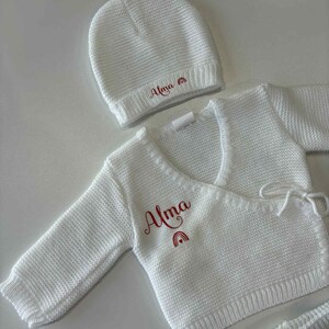 K&ouml;nnte beinhalten: Wei&szlig;es Strick-Baby-Set mit M&uuml;tze, Wickeloberteil und Shorts. Das Set ist mit dem Namen "Alma" in roter Schrift und einem Regenbogen-Design versehen. Der Stoff hat eine strukturierte Optik.