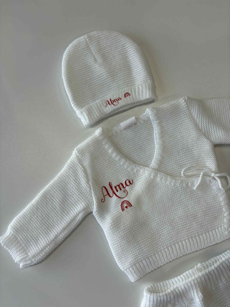 K&ouml;nnte beinhalten: Wei&szlig;es Strick-Baby-Set mit M&uuml;tze, Wickeloberteil und Shorts. Das Set ist mit dem Namen "Alma" in roter Schrift und einem Regenbogen-Design versehen. Der Stoff hat eine strukturierte Optik.