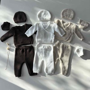 K&ouml;nnte beinhalten: Eine Auswahl an Babykleidung in neutralen Farbt&ouml;nen. Das Set umfasst Strick-Wickeljacken, Hosen, M&uuml;tzen und Babyschuhe. Die Farbpalette besteht aus Braun, Wei&szlig; und Beige, alles auf einer wei&szlig;en Oberfl&auml;che angeordnet.