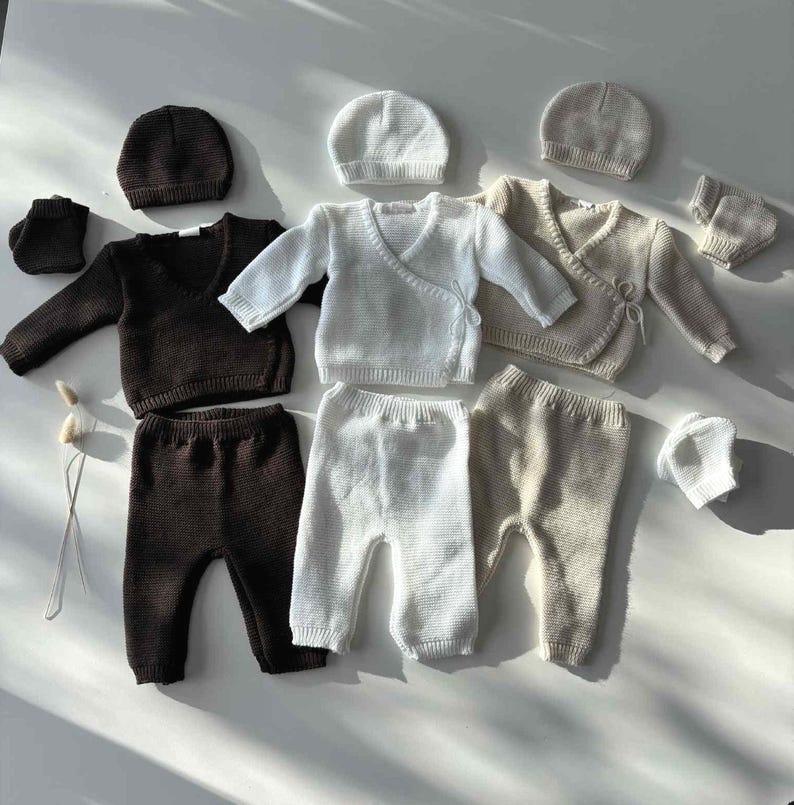 K&ouml;nnte beinhalten: Eine Auswahl an Babykleidung in neutralen Farbt&ouml;nen. Das Set umfasst Strick-Wickeljacken, Hosen, M&uuml;tzen und Babyschuhe. Die Farbpalette besteht aus Braun, Wei&szlig; und Beige, alles auf einer wei&szlig;en Oberfl&auml;che angeordnet.