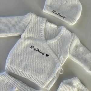 K&ouml;nnte beinhalten: Ein wei&szlig;es Strick-Baby-Outfit, bestehend aus einem Wickel-Cardigan, einer Hose, einer M&uuml;tze und F&auml;ustlingen. Der Name "Mathias" ist in schwarzer Schrift auf den Cardigan und die M&uuml;tze gestickt, mit einem kleinen schwarzen Herz auf dem Cardigan.