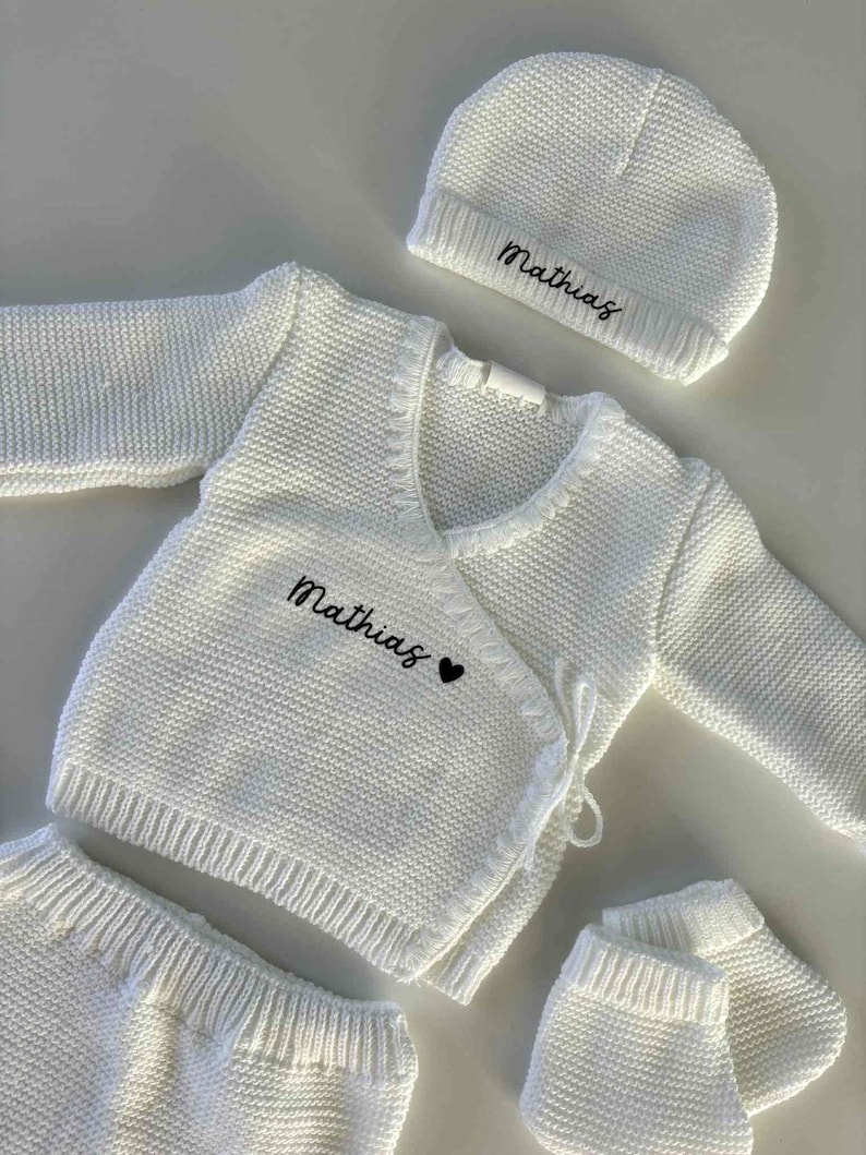 K&ouml;nnte beinhalten: Ein wei&szlig;es Strick-Baby-Outfit, bestehend aus einem Wickel-Cardigan, einer Hose, einer M&uuml;tze und F&auml;ustlingen. Der Name "Mathias" ist in schwarzer Schrift auf den Cardigan und die M&uuml;tze gestickt, mit einem kleinen schwarzen Herz auf dem Cardigan.