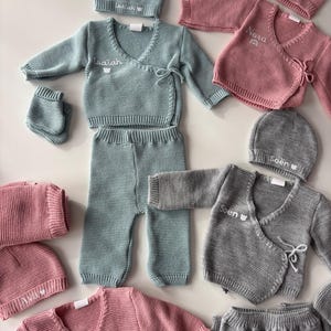 K&ouml;nnte beinhalten: Baby-Kleidungssets in verschiedenen Farben, darunter Hellblau, Rosa und Grau. Jedes Set enth&auml;lt eine M&uuml;tze, ein Wickeloberteil, eine Hose und F&auml;ustlinge. Die Namen "Isaiah", "Nora", "Soen" und "Ava" sind auf den Kleidungsst&uuml;cken gestickt.