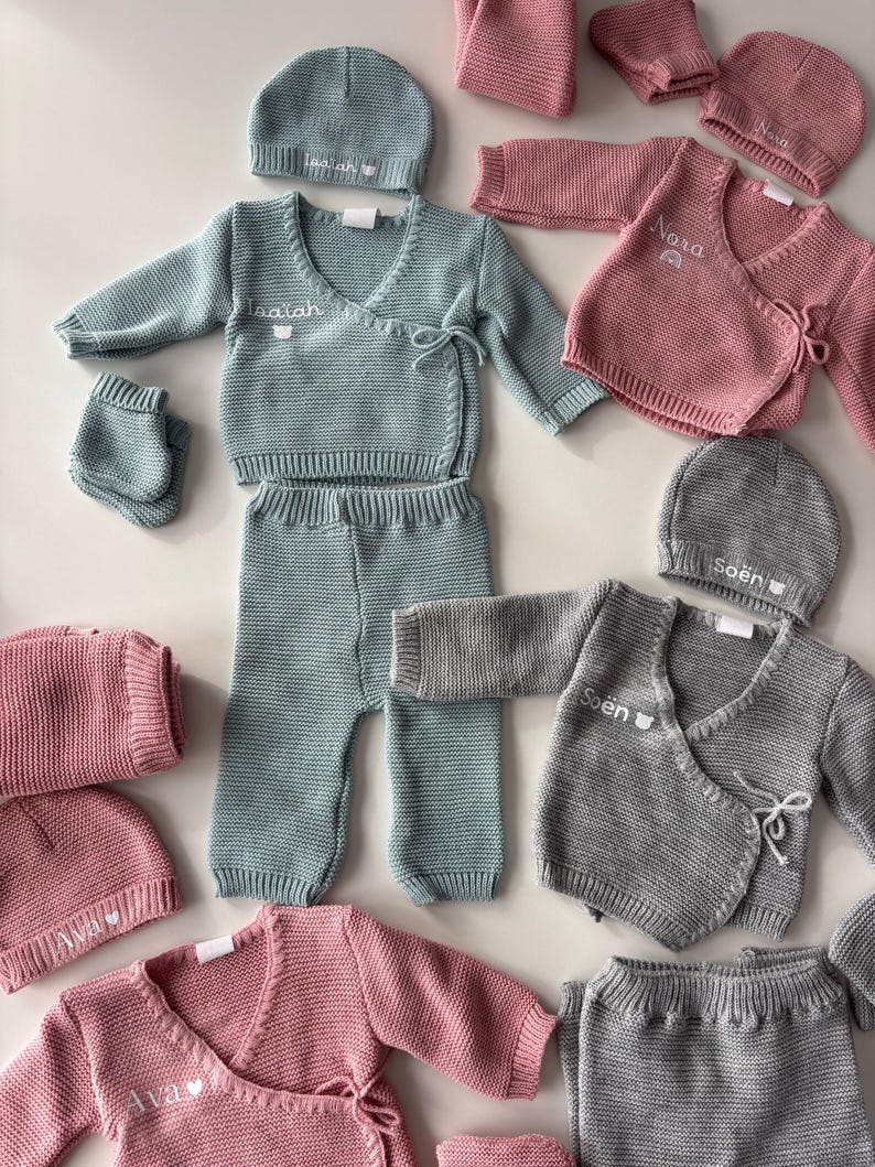 K&ouml;nnte beinhalten: Baby-Kleidungssets in verschiedenen Farben, darunter Hellblau, Rosa und Grau. Jedes Set enth&auml;lt eine M&uuml;tze, ein Wickeloberteil, eine Hose und F&auml;ustlinge. Die Namen "Isaiah", "Nora", "Soen" und "Ava" sind auf den Kleidungsst&uuml;cken gestickt.
