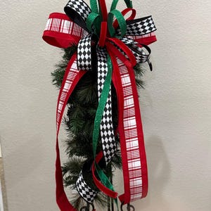 May include: A Christmas tree topper featuring a festive arrangement of red, green, black, and white ribbons. The ribbons include a red and white plaid pattern, black and white diamond pattern, and solid red and green ribbons. The topper is attached to a small artificial tree.