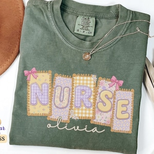 Peut inclure: T-shirt vert mousse avec le mot "NURSE" dans un motif patchwork avec des motifs floraux et vichy. Le nom "olivia" est écrit en dessous. Un collier doré et un chapeau marron sont également visibles.