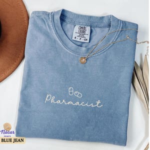May include: A blue jean Comfort Colors t-shirt with the word "Pharmacist" and two pill graphics in white. A gold necklace with a pendant is on the shirt. The shirt has a small tag that says "Comfort Colors". The bottom left corner of the image has the text "Nacar Prints BLUE JEAN".