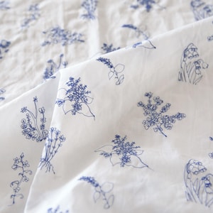 Pode incluir: Tecido branco com um padrão repetido de bordados florais azuis. As flores bordadas incluem raminhos, cachos e flores individuais. O tecido está ligeiramente enrugado, sugerindo que é um material macio, possivelmente algodão ou linho.