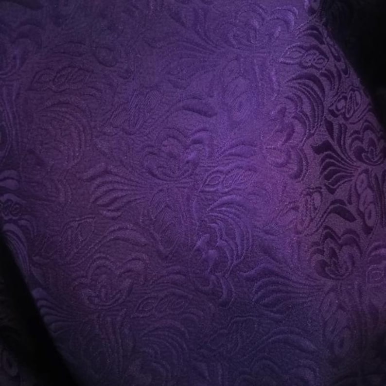 May include: Purple fabric with a textured, floral pattern. The fabric has a subtle sheen and a soft, luxurious feel.