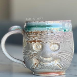Puede incluir: Una taza de cerámica con un diseño pintado a mano de la cara de un gato. La taza está esmaltada en un color gris claro con acentos marrones y verdes. La cara del gato está esculpida con una amplia sonrisa y ojos grandes. La taza tiene un asa y está hecha por ESH Pottery.