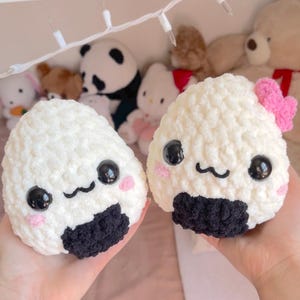 May include: Two handmade, white crochet onigiri plushies with black nori details, black eyes, pink cheeks, and smiling faces. One has a pink bow. The background features stuffed animals and fairy lights.