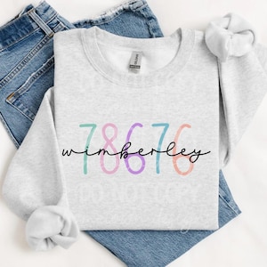 Op de afbeelding: Een lichtgrijs sweatshirt met de tekst "7867 Wimberley" gedrukt op de voorkant in kleurrijke, gestileerde cijfers.