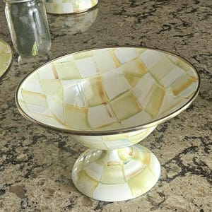 May include: A decorative pedestal bowl with a checkered pattern in shades of white, yellow, and brown. The bowl has a silver-colored rim and a matching pedestal base. A glass jar with a silver lid is visible in the background.