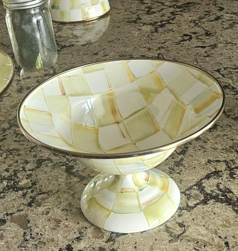 May include: A decorative pedestal bowl with a checkered pattern in shades of white, yellow, and brown. The bowl has a silver-colored rim and a matching pedestal base. A glass jar with a silver lid is visible in the background.