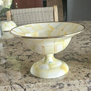 May include: A decorative pedestal bowl with a wide, shallow top and a rounded base. The bowl features a checkered pattern in shades of yellow, green, and white, with a gold-colored rim. The bowl is likely made of ceramic or porcelain.
