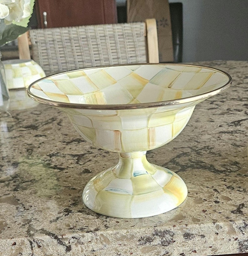 May include: A decorative pedestal bowl with a wide, shallow top and a rounded base. The bowl features a checkered pattern in shades of yellow, green, and white, with a gold-colored rim. The bowl is likely made of ceramic or porcelain.
