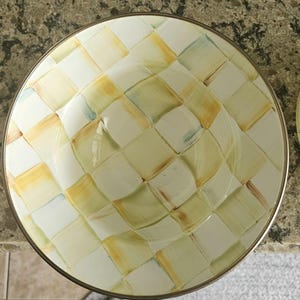 May include: Overhead view of a round, decorative bowl with a gold rim. The bowl features a checkered pattern in shades of yellow, green, and white. The design is reminiscent of a woven basket or fabric.
