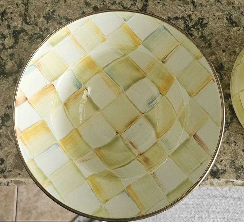 May include: Overhead view of a round, decorative bowl with a gold rim. The bowl features a checkered pattern in shades of yellow, green, and white. The design is reminiscent of a woven basket or fabric.