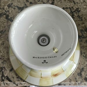 May include: White and yellow checkered ceramic pedestal bowl with a wide rim and a flared base. The bowl has a metal screw in the center and the words "MacKenzie-Childs" are printed on the side.
