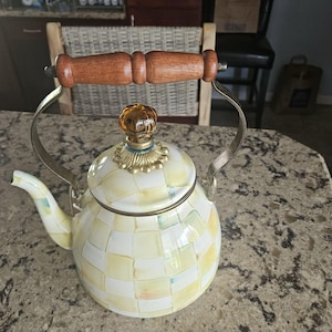 Puede incluir: Una tetera decorativa con un patrón a cuadros en tonos amarillos y blancos. Cuenta con un asa de madera, un adorno de tapa dorado y detalles metálicos plateados. La tetera está diseñada para su uso en la estufa.