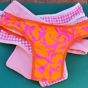 May include: Three pairs of bikini bottoms in a stack. The top pair is patterned with orange and pink florals, the middle pair is pink gingham, and the bottom pair is plain pink. All have orange edging.