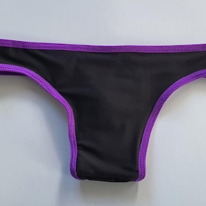 Puede incluir: Ropa interior negra con ribete morado. La prenda tiene un corte de cintura baja y está hecha de una tela lisa y sólida. Los bordes están acabados con un ribete morado en contraste, agregando un toque de color al diseño.