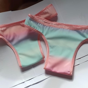 May include: Two pairs of pastel-coloured underwear. The fabric transitions from pink to blue-green, with a pink trim. The underwear is laid out on a white surface. The style is a thong.